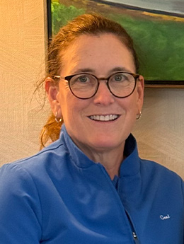 The image shows a person wearing glasses, smiling at the camera, with short brown hair and dressed in a blue jacket.