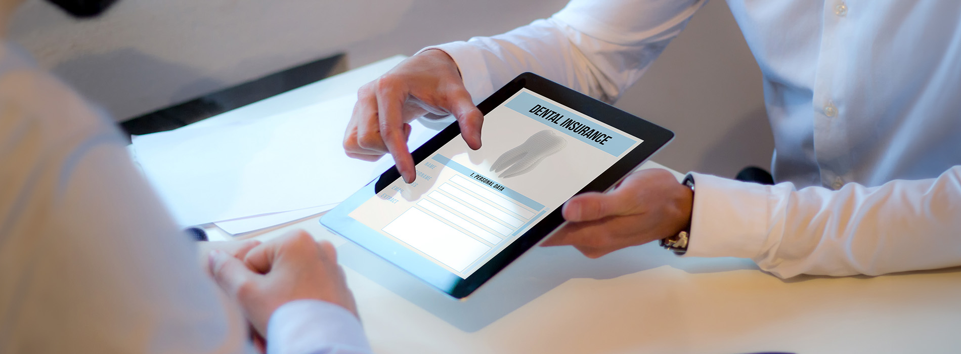 The image shows two hands holding a tablet with an open document on a table, in front of which there are two people engaged in conversation.