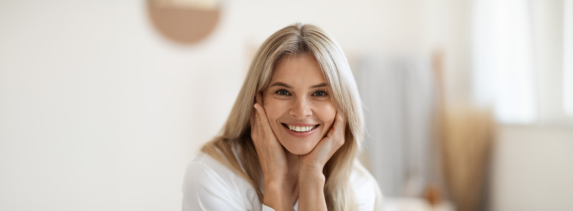 A woman with blonde hair smiling at the camera, wearing a white top, with her hand on her face.