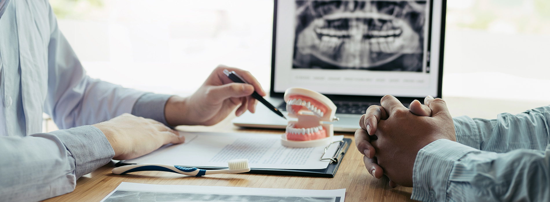 Two people sitting at a table with open laptops, one person writing on paper, both wearing masks, and one looking at medical images on a screen behind them.