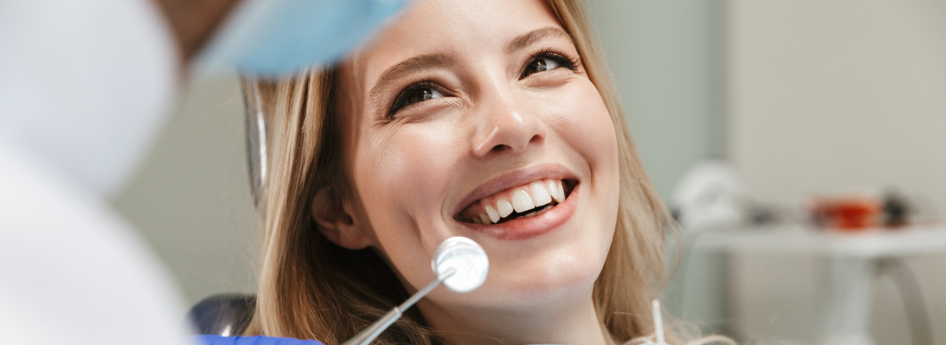The image depicts a young woman with a bright smile, sitting in a dental chair with a dentist s hand visible in the foreground, holding a dental tool near her mouth.