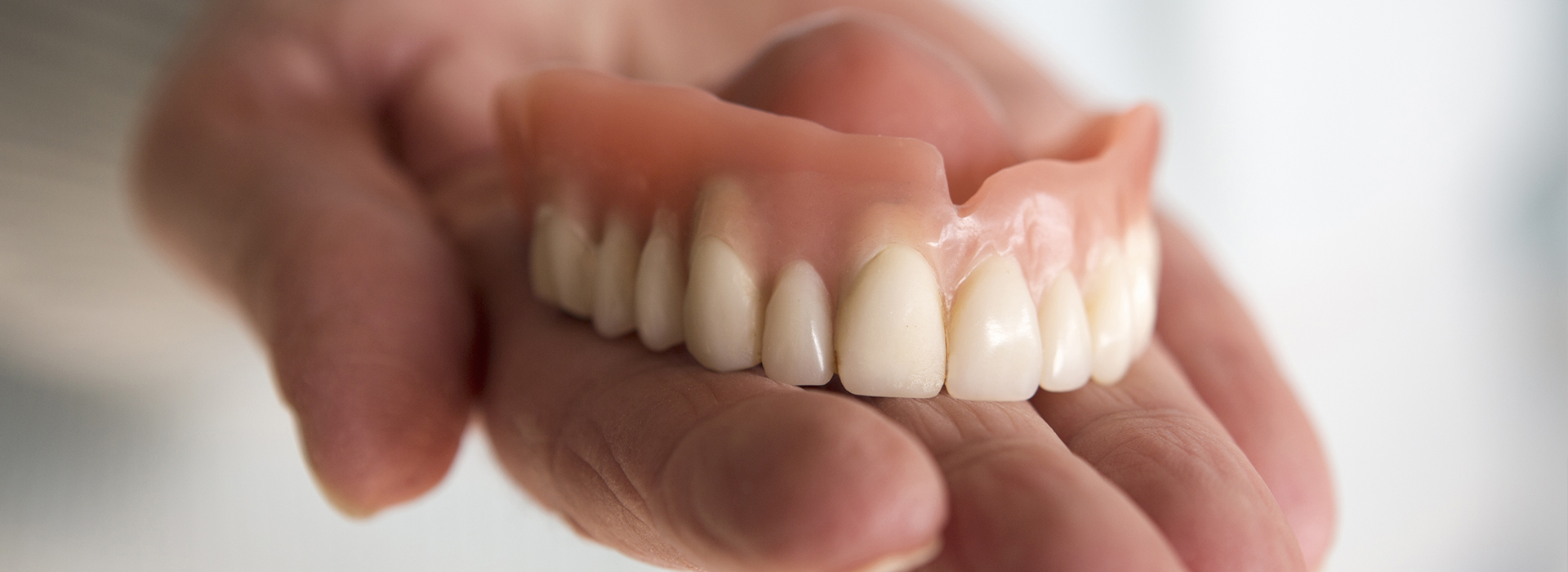 The image shows a hand holding an open mouth with fake teeth, possibly for dental implant purposes, against a blurred background.